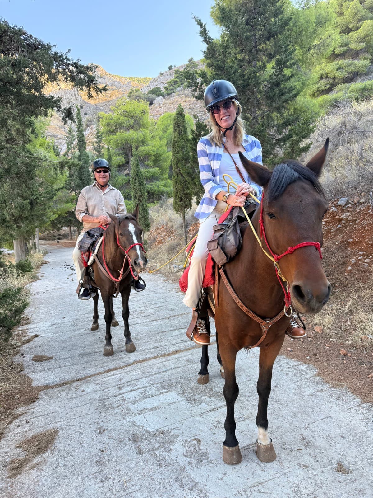 Two smiling riders on horseback on a pine-lined path above Hydra