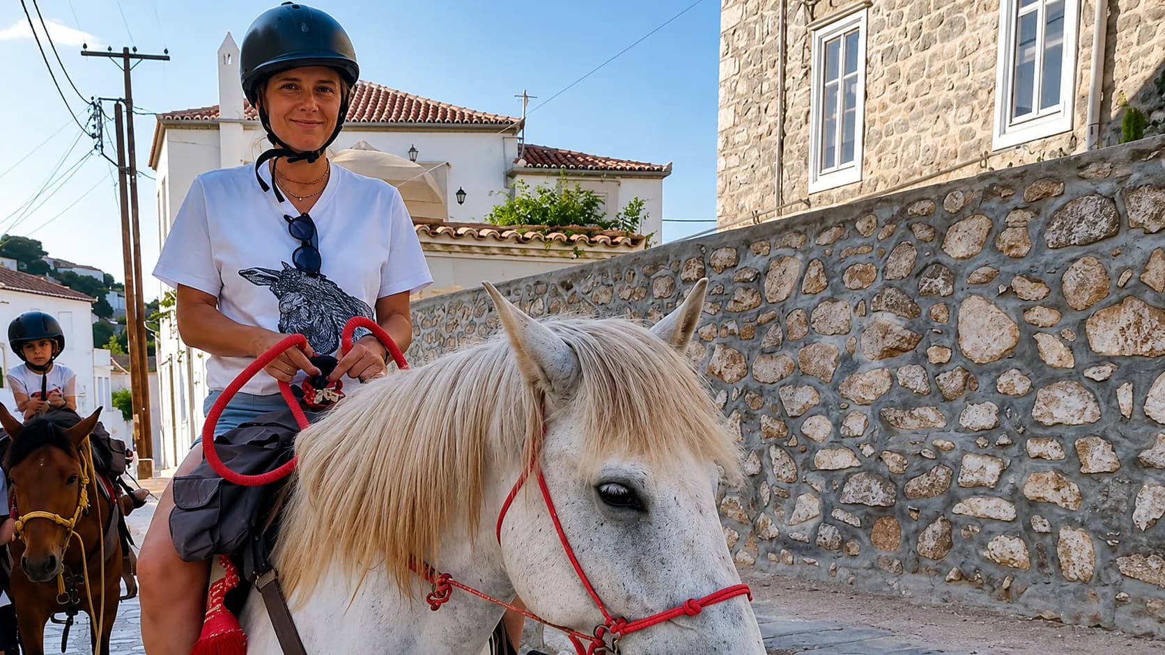 A child in a riding helmet on a pony behind an adult on a white horse, on a stone lane in Hydra town