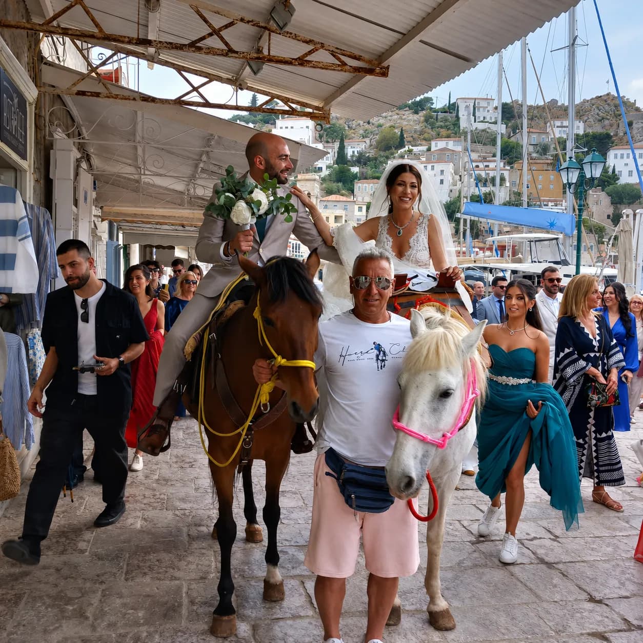 A bride and groom arriving on horseback along the Hydra waterfront, wedding guests following behind